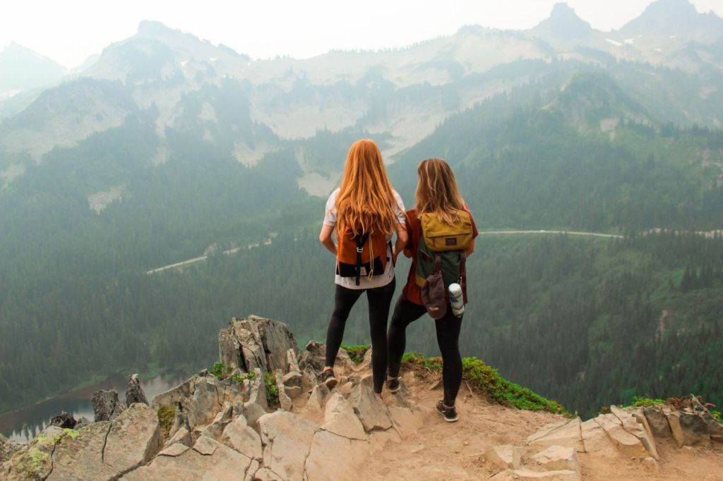 Two woman wearing backpacks standing ont he edge of a cliff looking out at a valley and mountains in the distance
