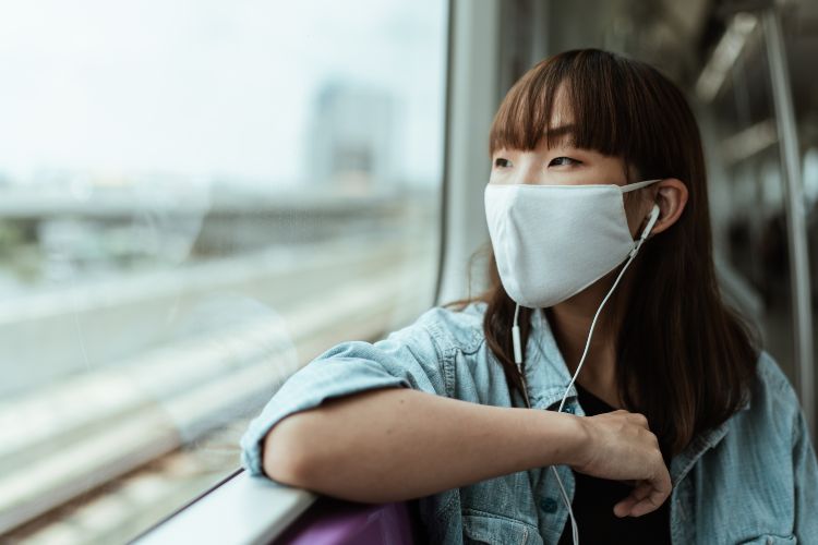 A woman wearing a white mask and ear buds looking out of the window of a moving train